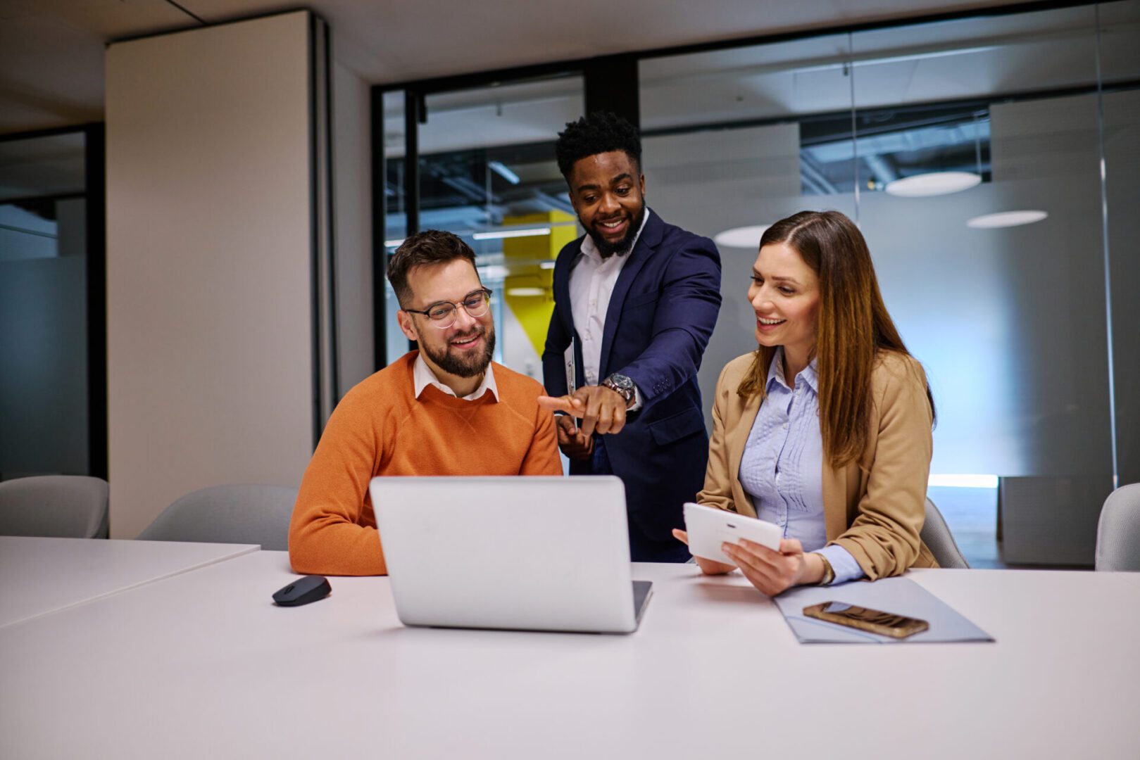Three businesspeople are collaborating on a project, using a laptop and a digital tablet in a modern office, discussing strategies and sharing ideas