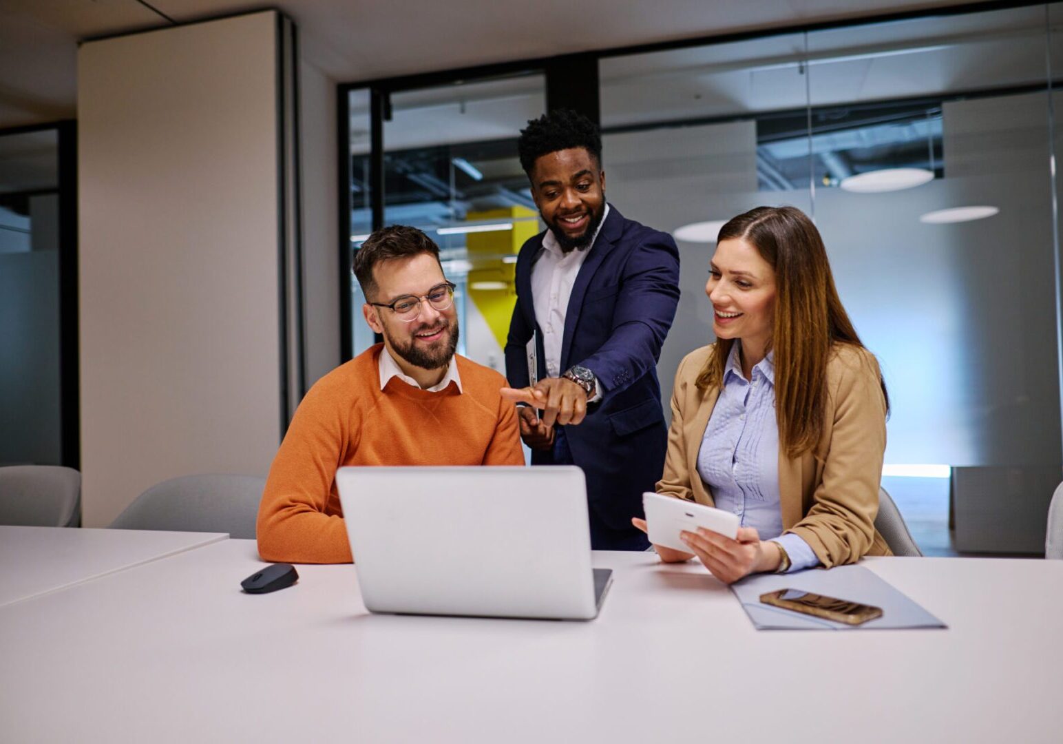 Three businesspeople are collaborating on a project, using a laptop and a digital tablet in a modern office, discussing strategies and sharing ideas